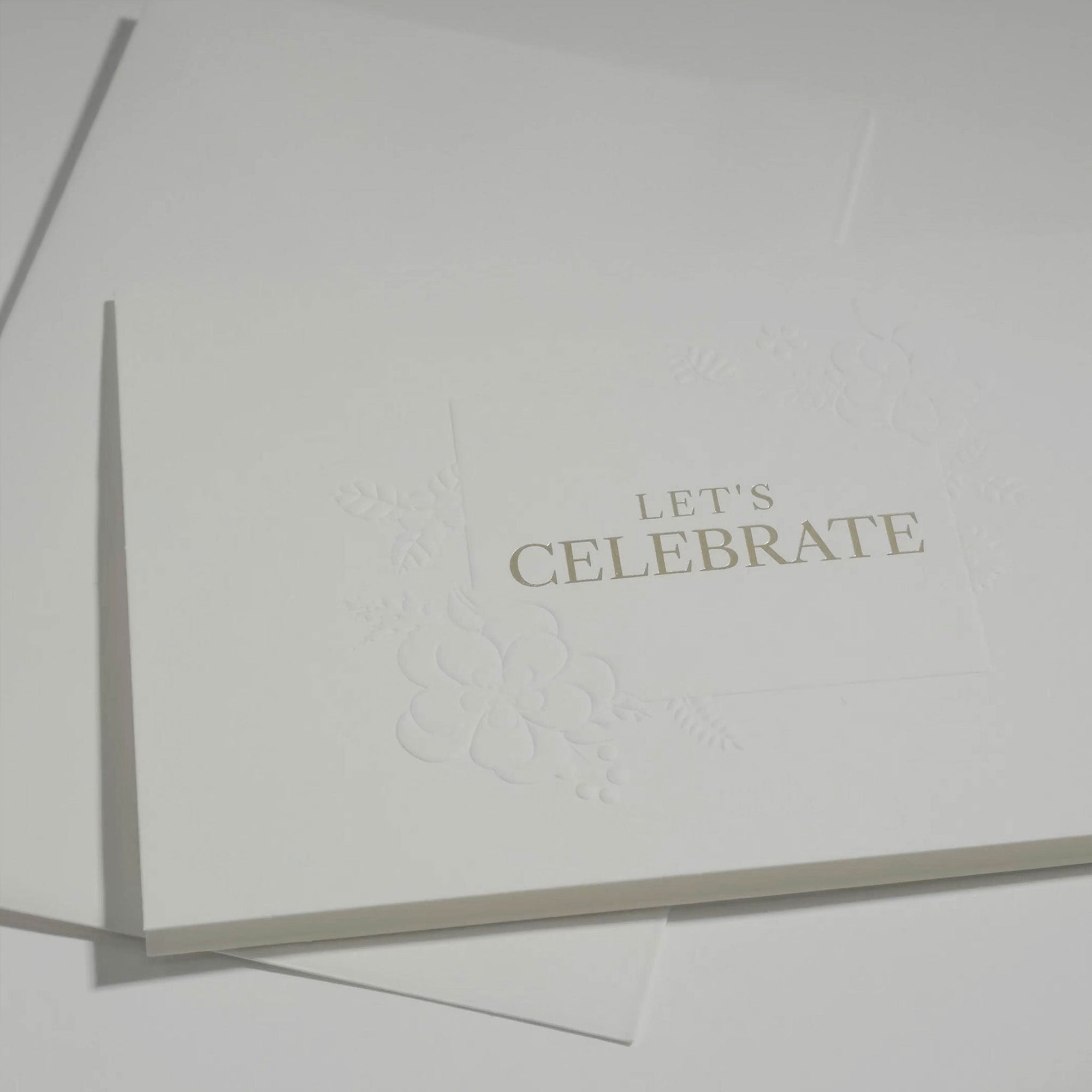 Angled top-down shot of the premium minimalist white celebration note card highlighting its crisp gold lettering and fine embossed border details.