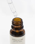 A clear glass pipette dropper dispensing a liquid droplet into an open amber bottle of gardenia essential oil.