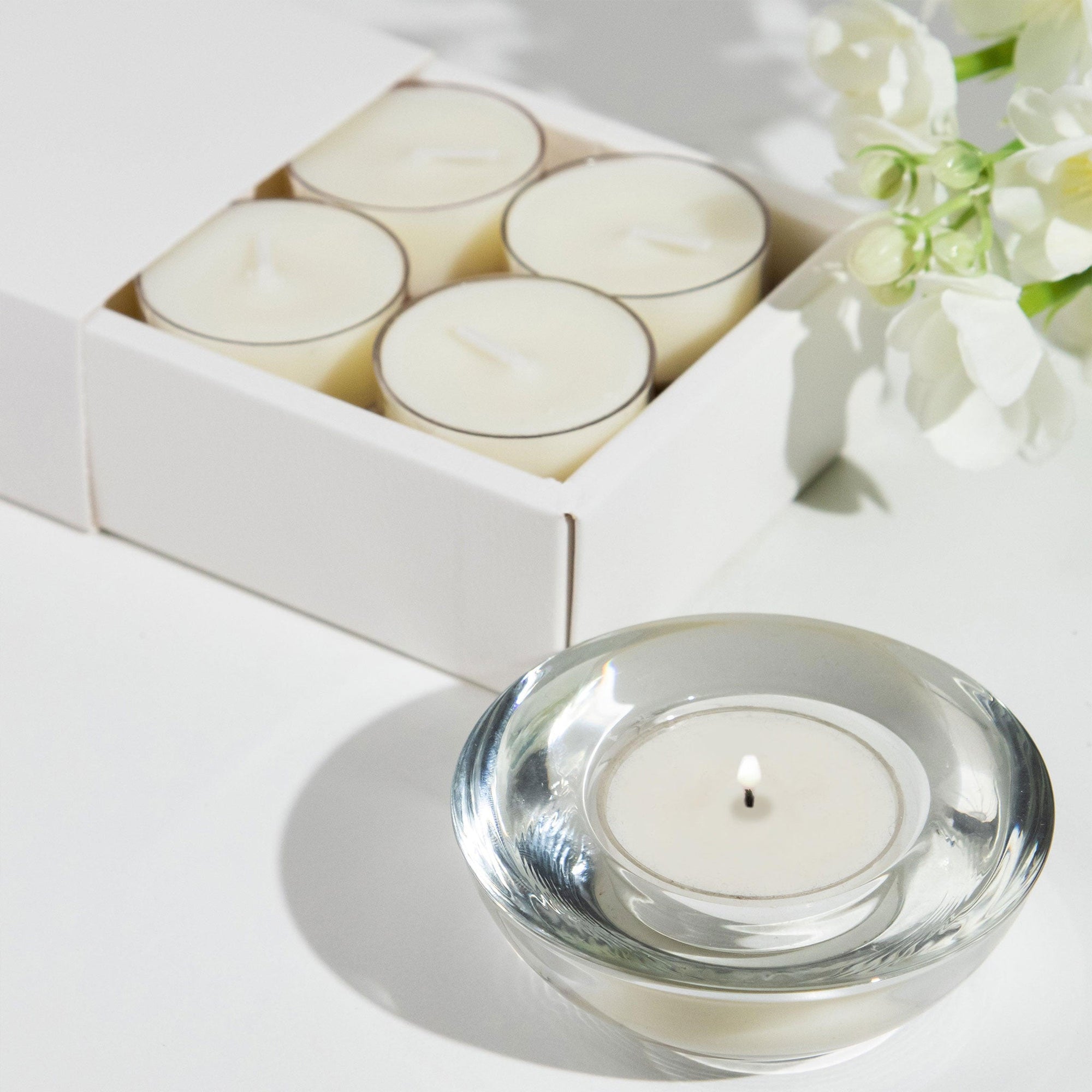 A lit Vaucluse Jasmine tealight candle burning inside a clear glass holder, placed in front of a white product box and delicate white flowers.