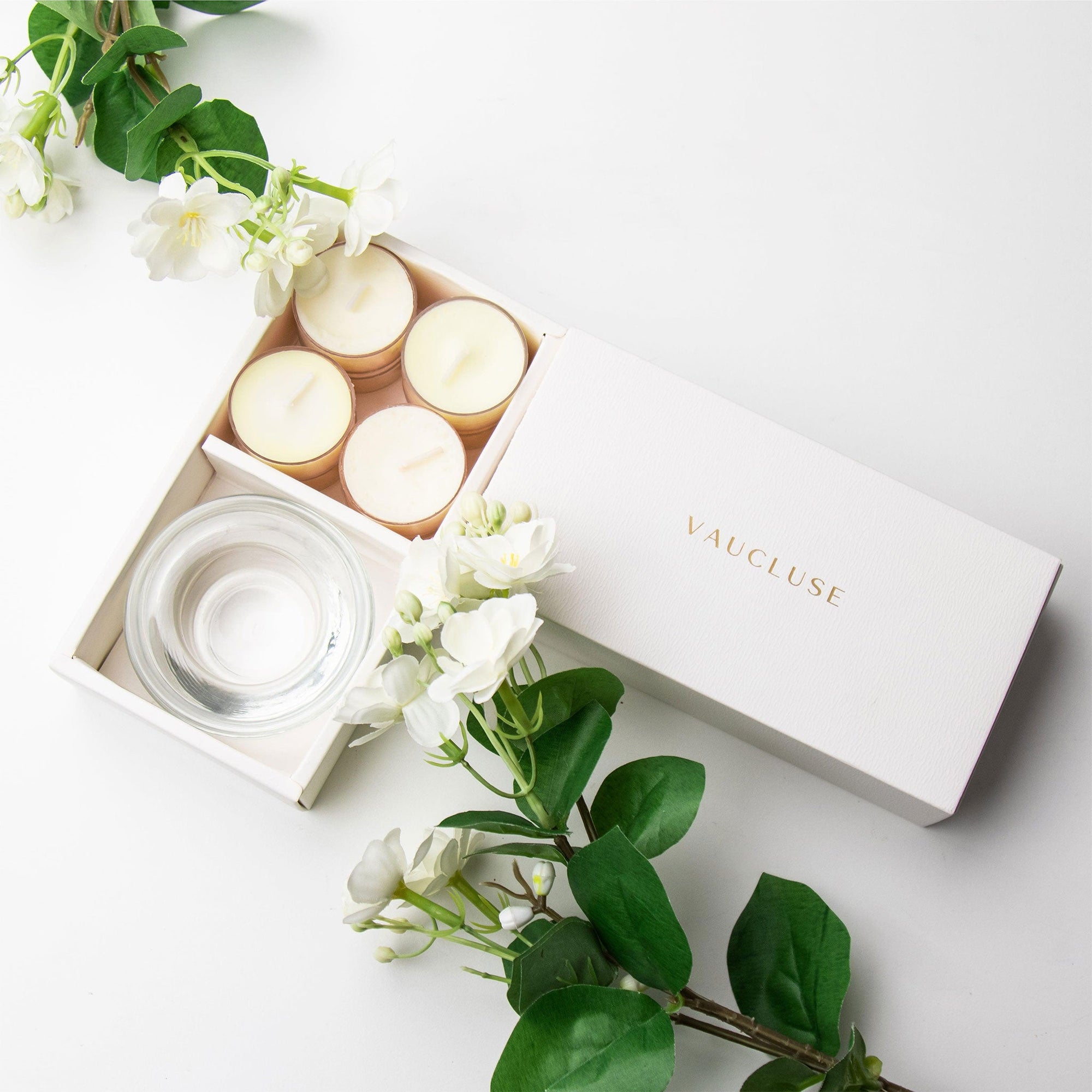Top-down minimalist flat lay of the open jasmine tealight candle set styled beautifully with fresh white jasmine flowers and green leaves on a white surface.