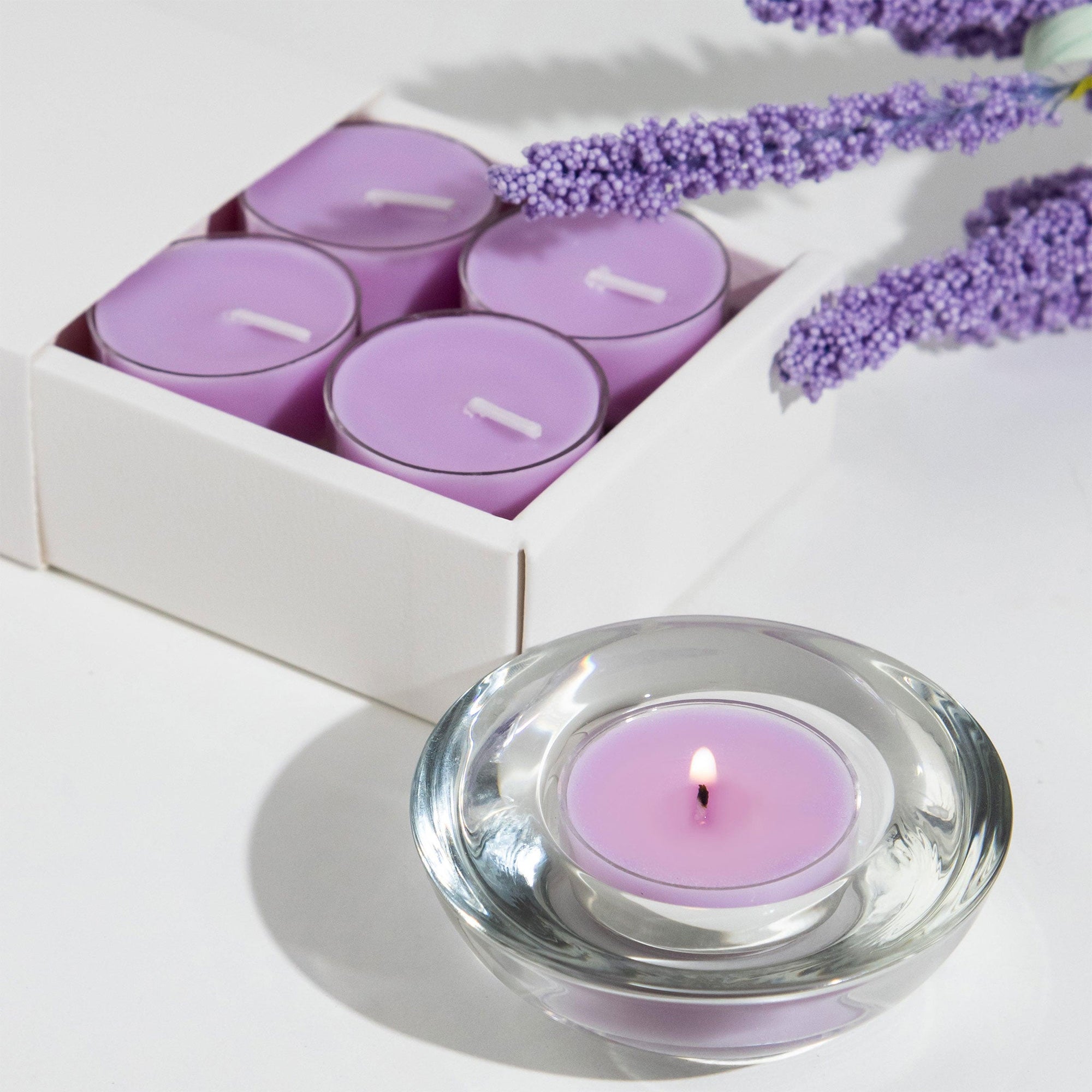 A lit purple Vaucluse Lavender tealight candle burning in a clear glass holder, styled with a white product box and a sprig of dried lavender flowers.