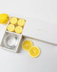 Minimalist overhead flat lay of the open yellow tealight candle set styled with a whole fresh lemon and two vibrant citrus slices on a white background.
