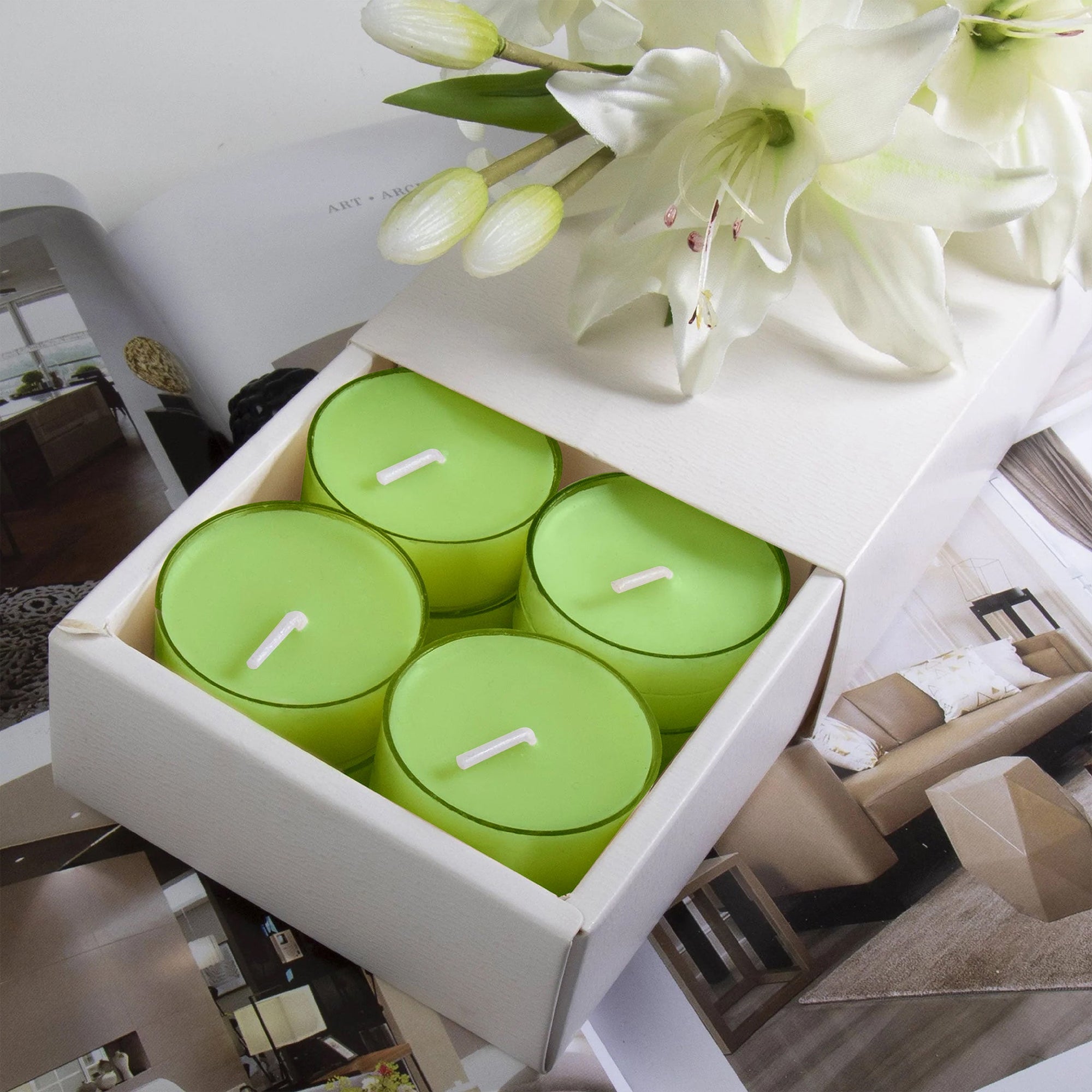 A premium white Vaucluse 16-pack box sliding open to reveal the top tier of light green lily tealight candles, elegantly styled below fresh white lilies.