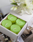 A premium white Vaucluse 16-pack box sliding open to reveal the top tier of light green lily tealight candles, elegantly styled below fresh white lilies.