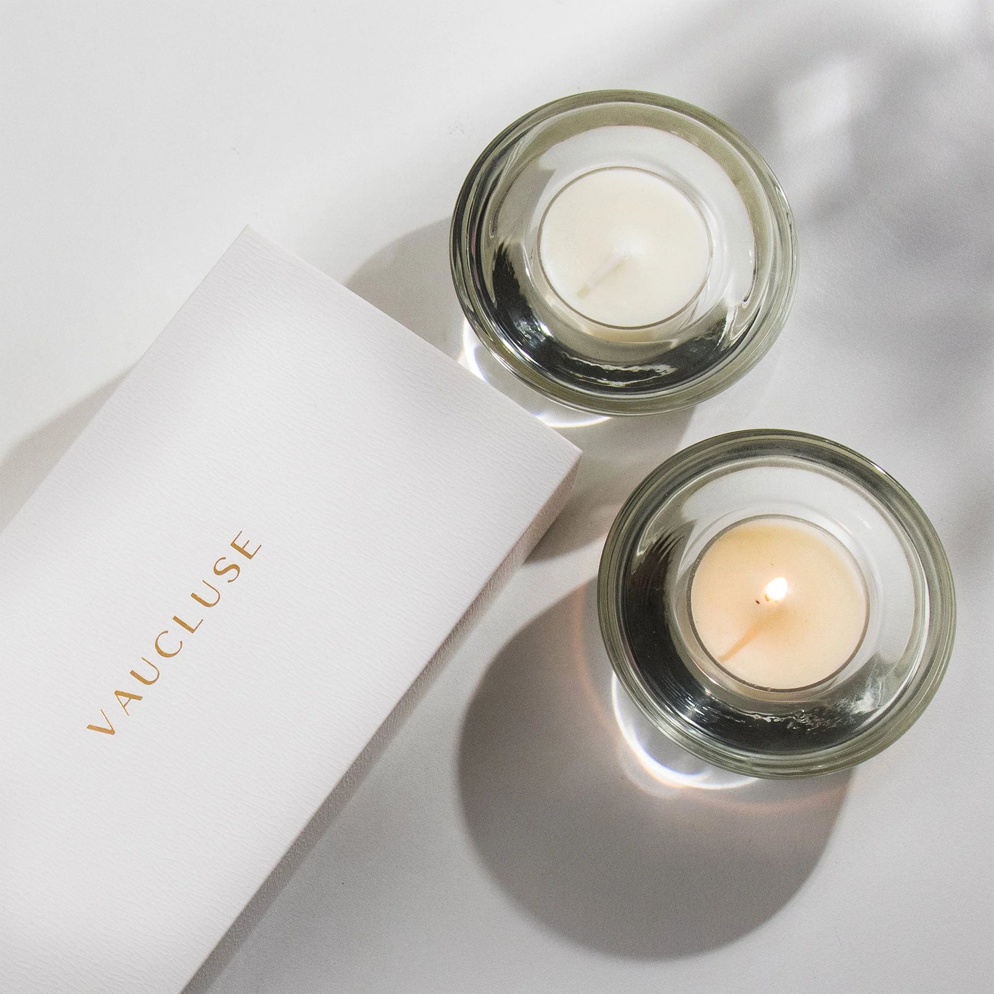 An overhead top-down view of a lit vanilla scented tealight candle softly burning in a clear glass holder alongside a white Vaucluse luxury gift box.