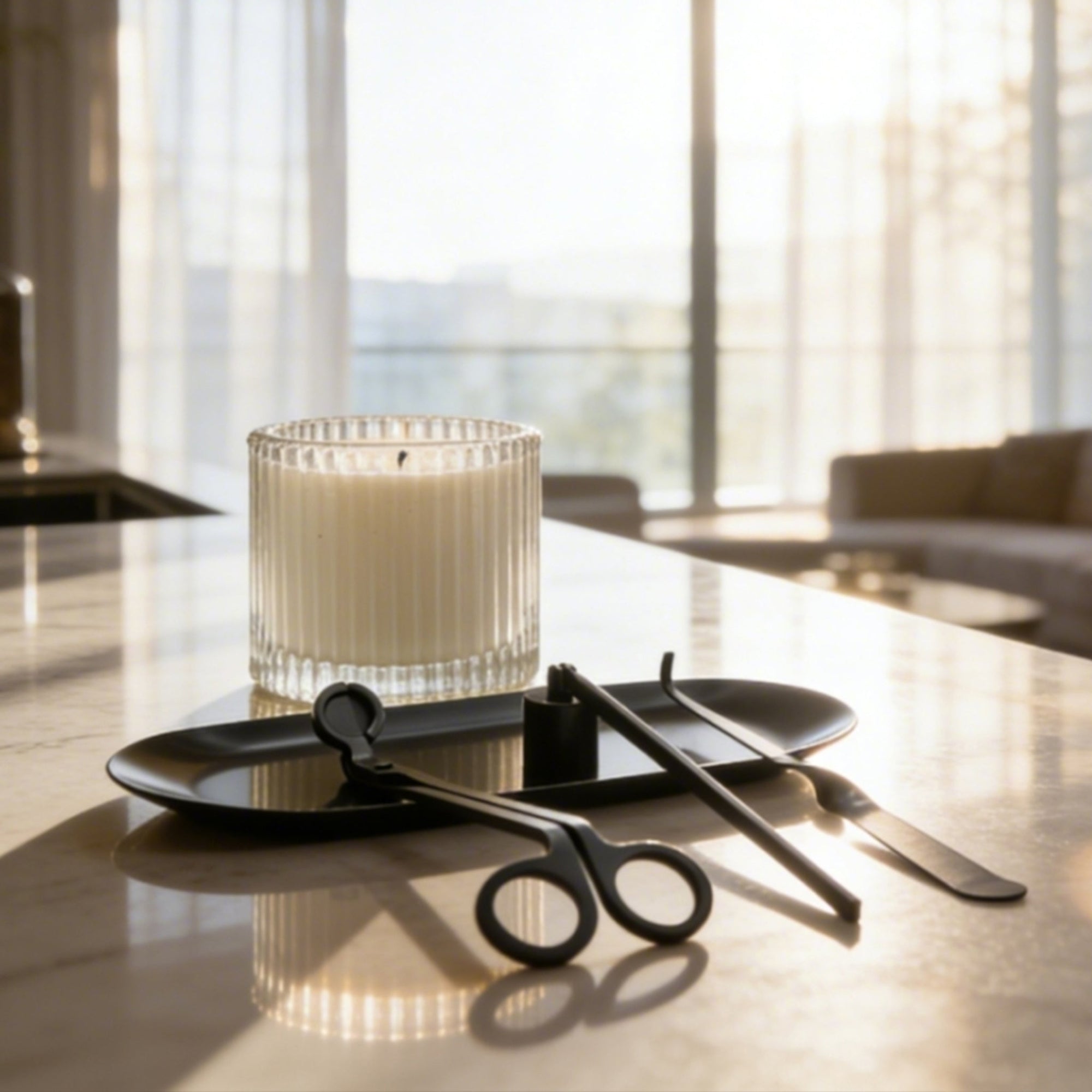 Complete matte black candle care set with a trimmer, snuffer, and dipper arranged on a tray next to a lit ribbed glass candle in a bright modern room.