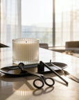 Complete matte black candle care set with a trimmer, snuffer, and dipper arranged on a tray next to a lit ribbed glass candle in a bright modern room.