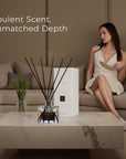 An opulent Vaucluse black rose reed diffuser showcased on a modern marble coffee table, with a woman in an elegant dress sitting on a beige couch in the background.