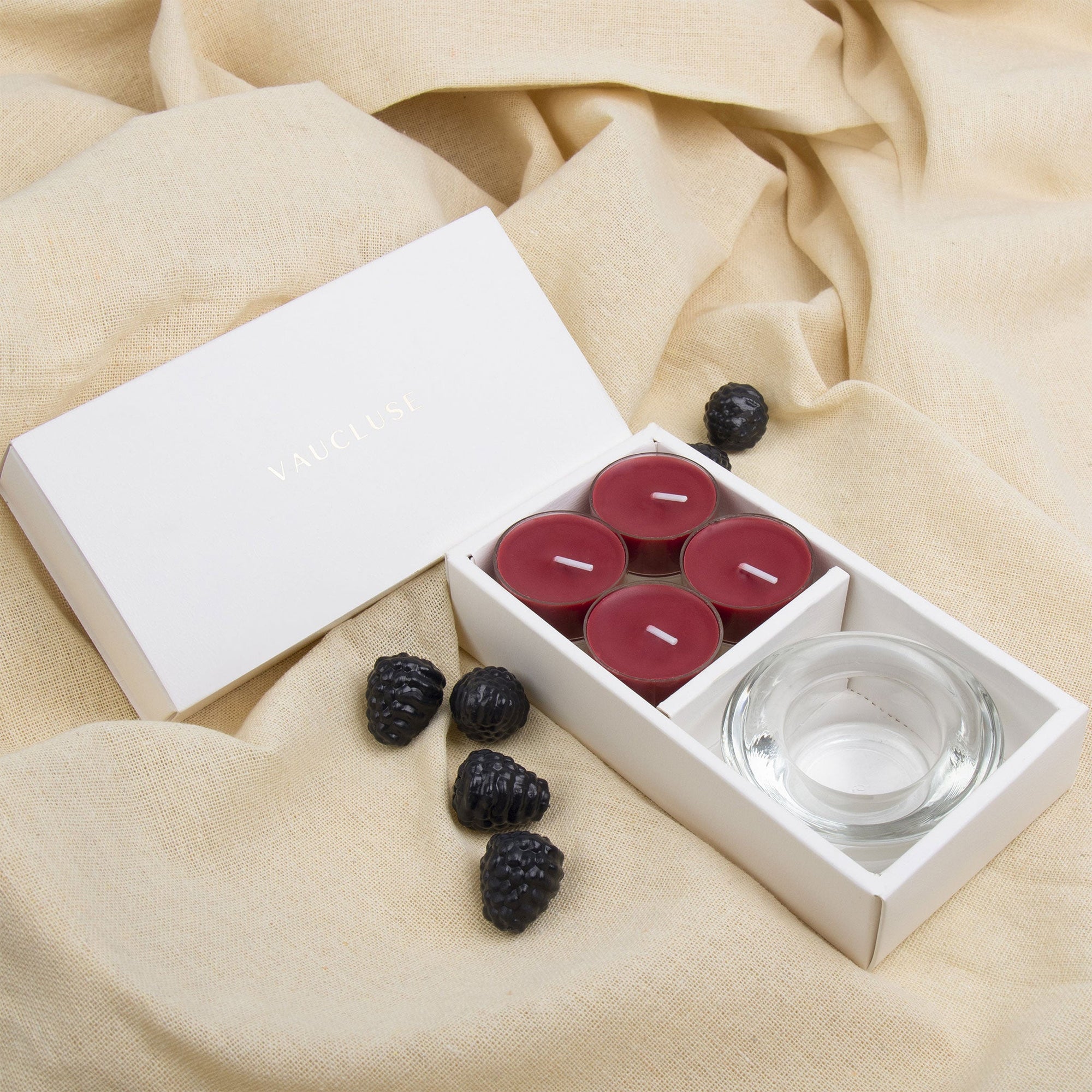 Elegant lifestyle scene showing the deep plum blackberry tealight aromatherapy gift set styled on draped beige linen fabric with fresh blackberry accents.