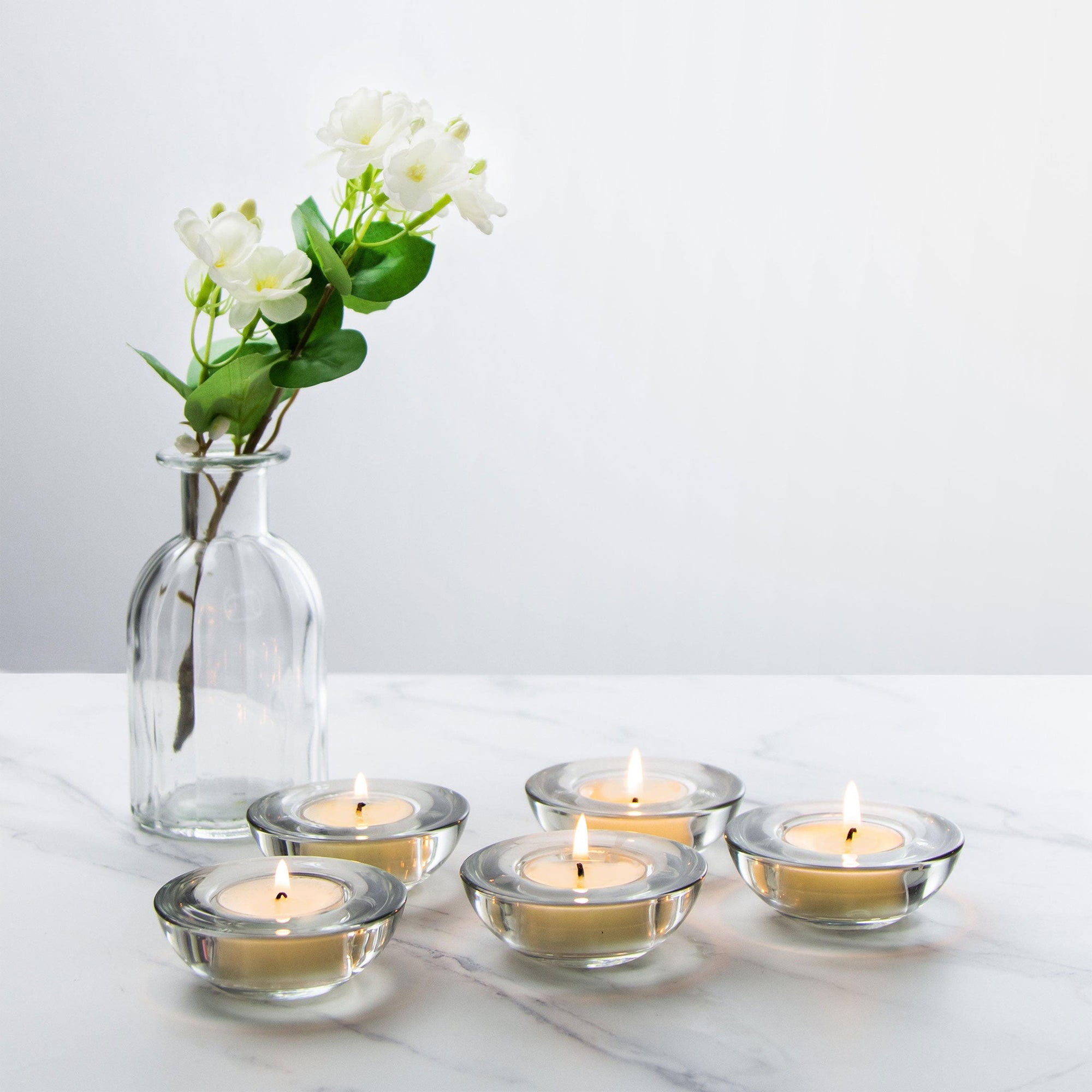 Five premium clear glass tealight holders with lit warm candles arranged elegantly on a white marble surface next to a clear glass flower vase.