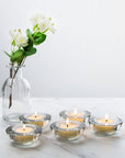 Five premium clear glass tealight holders with lit warm candles arranged elegantly on a white marble surface next to a clear glass flower vase.