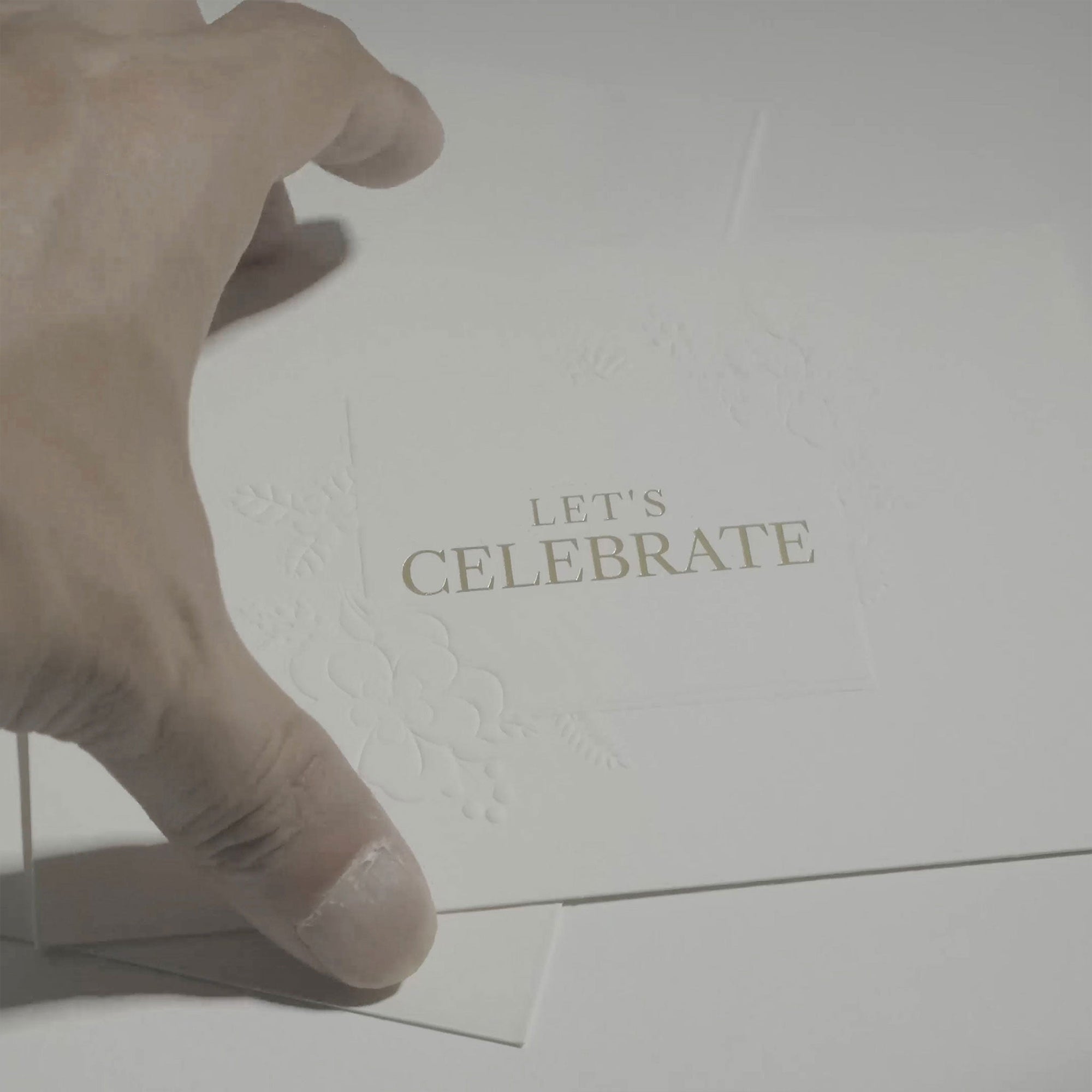 A close-up view of a person's hand interacting with the elegant white Let's Celebrate greeting card against a clean, minimalist background.