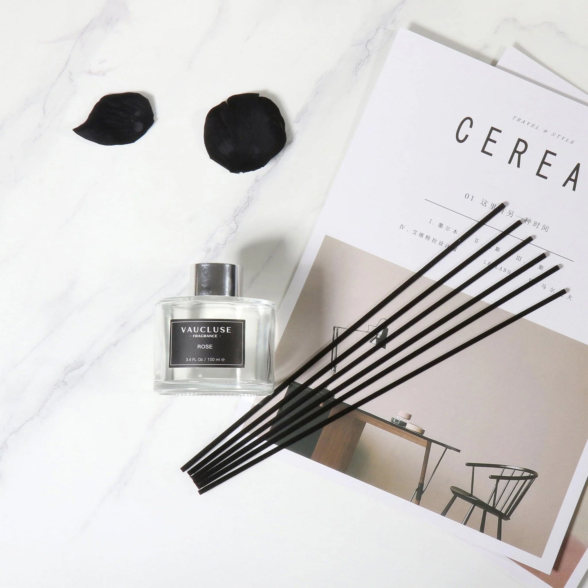 An overhead flat lay view of a clear glass Vaucluse reed diffuser bottle with a cluster of black reeds arranged on a stylized magazine with dark rose petals on a marble counter.