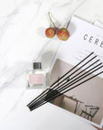An overhead flat lay view of a clear glass Vaucluse diffuser bottle and its black reeds arranged on an aesthetic magazine alongside fresh lychee fruits on a marble surface.
