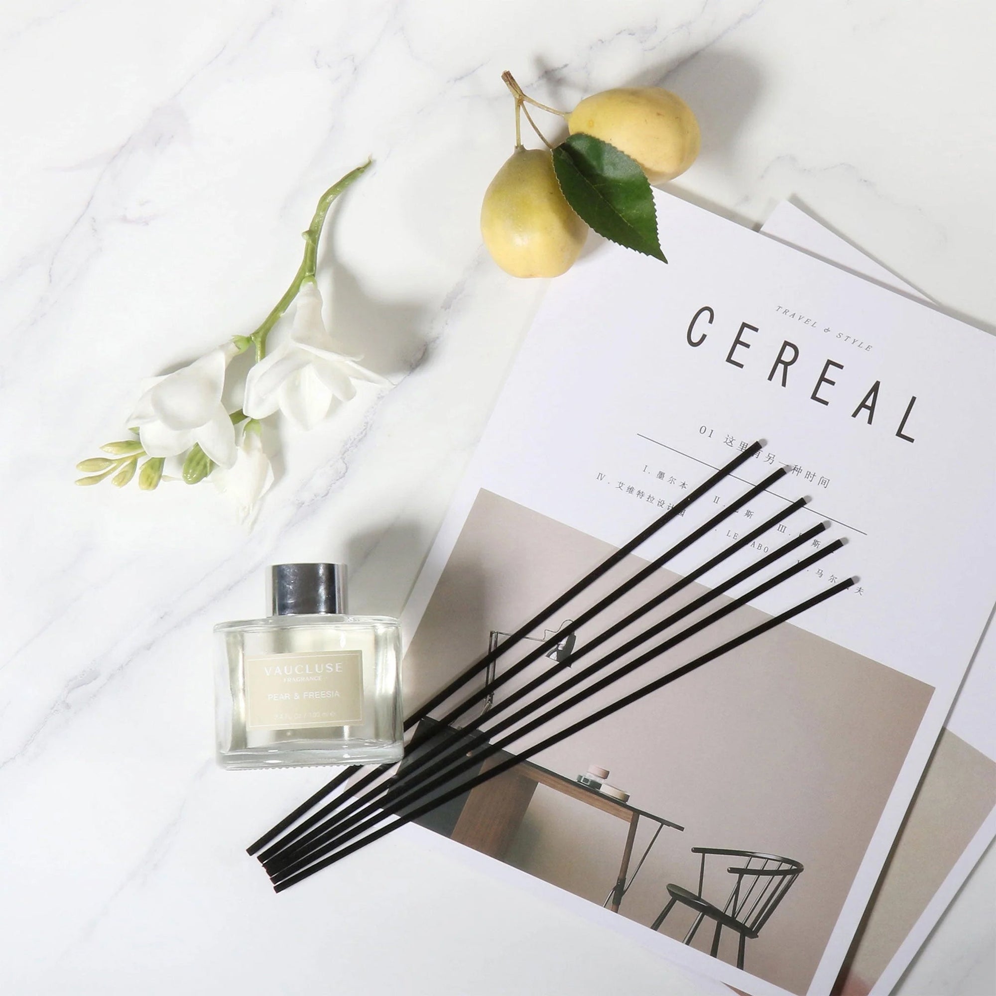 An overhead flat lay view of a clear glass Vaucluse diffuser bottle with black reeds arranged on a magazine next to white flowers and fresh green pears on a marble surface.