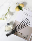 An overhead flat lay view of a clear glass Vaucluse diffuser bottle with black reeds arranged on a magazine next to white flowers and fresh green pears on a marble surface.