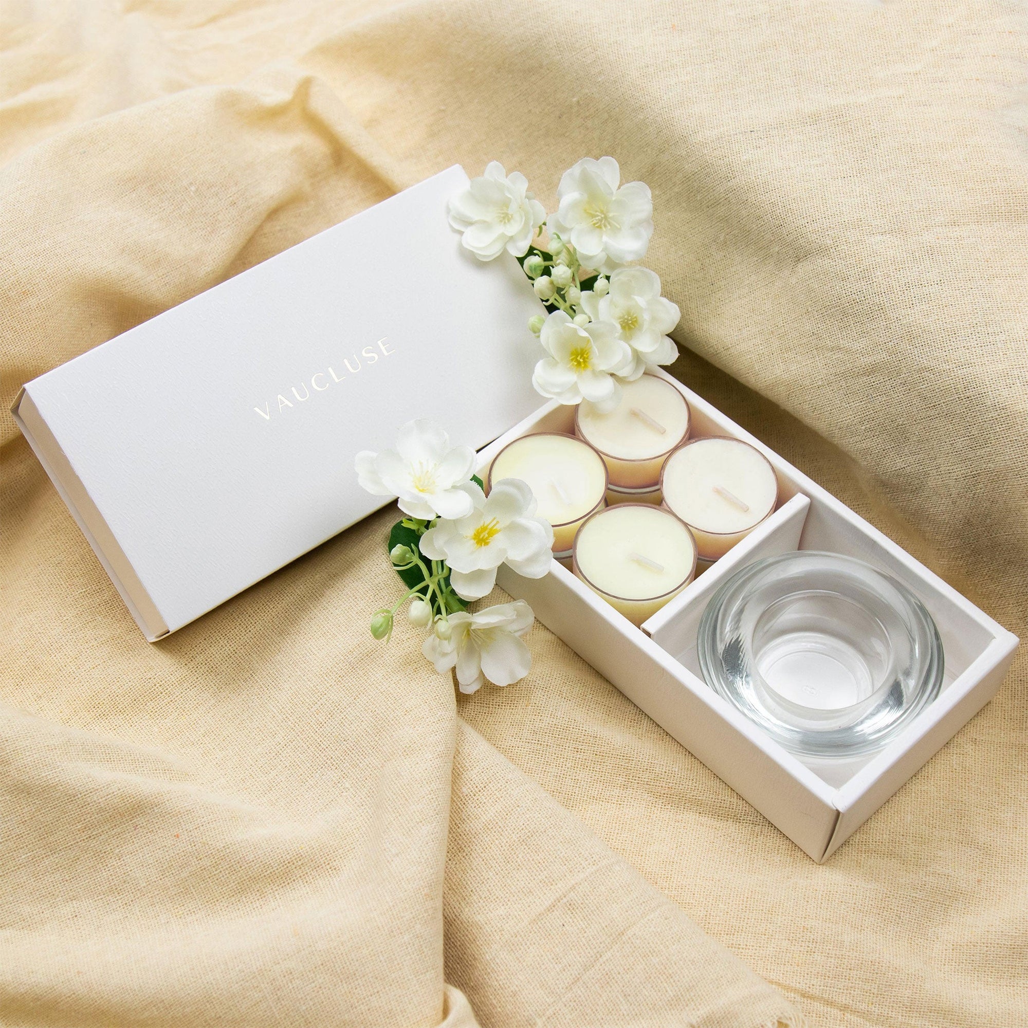 Cozy lifestyle display featuring the open Vaucluse jasmine tealight set resting on draped beige linen fabric decorated with white jasmine blossoms.