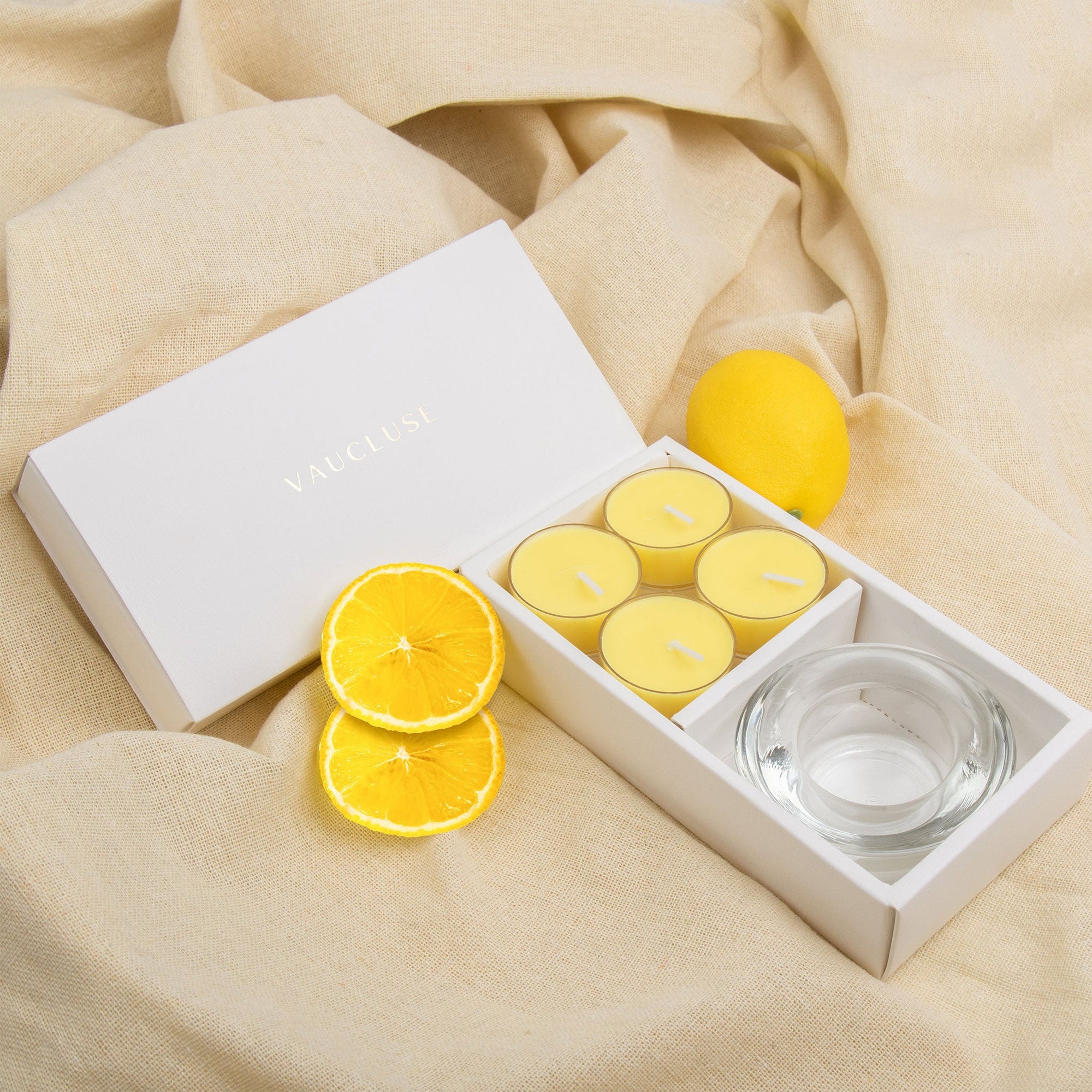 Elegant lifestyle scene showing the lemon tealight aromatherapy gift set styled on draped beige linen fabric with fresh lemon accents.