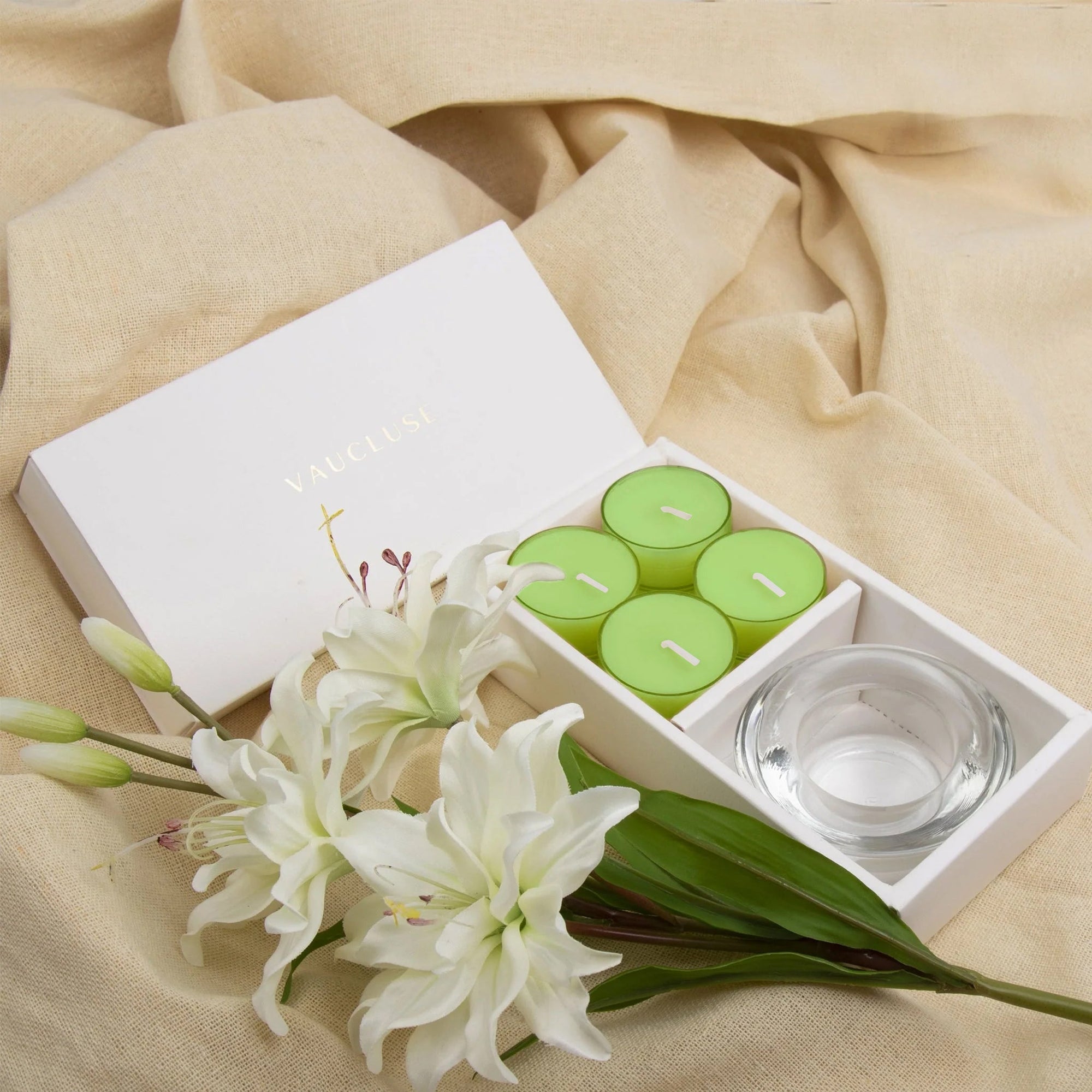Elegant lifestyle layout showing the green lily tealight set and clear glass holder nestled on a soft, draped beige linen fabric with fresh white lilies.