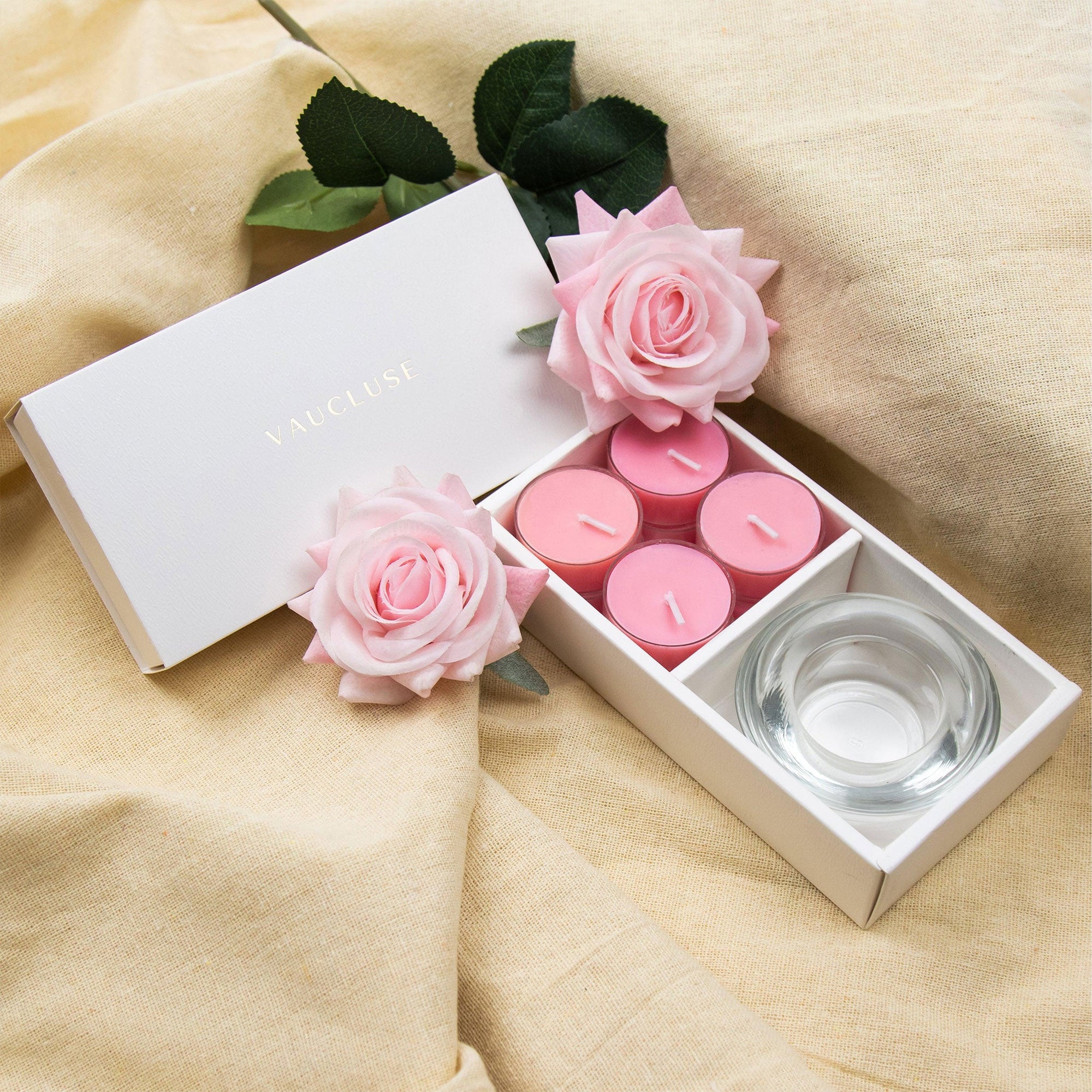 Elegant aromatherapy lifestyle scene featuring the open rose tealight gift box styled on draped beige linen fabric with light pink decorative roses.