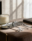 An elegant silver candle care set arranged on its matching tray resting on a plush brown velvet ottoman next to a ribbed candle.