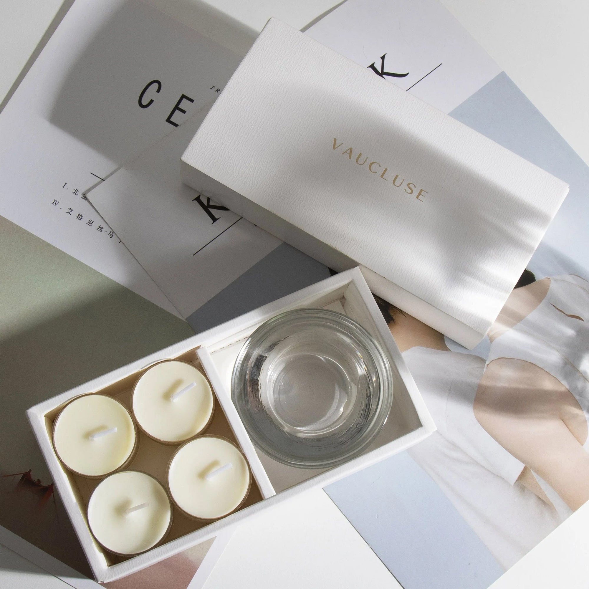 Luxury vanilla scented tealights and clear glass holder arranged in an open branded white box over chic editorial design books.