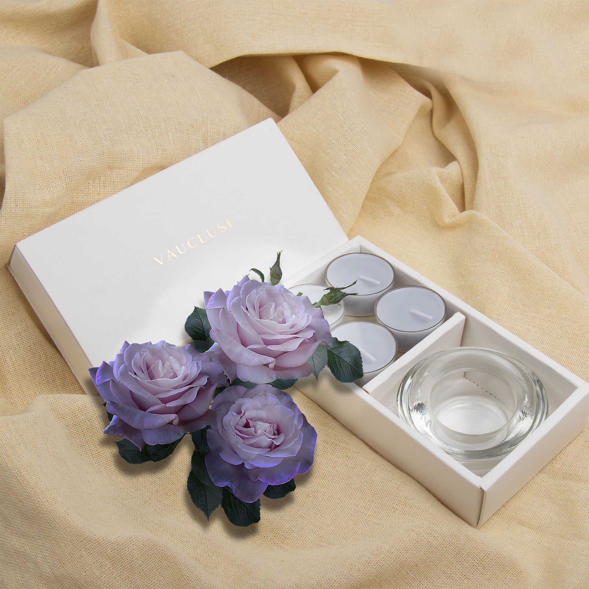 Elegant lifestyle scene featuring the open velvet tealight gift set styled on draped beige linen fabric with decorative purple roses.