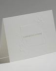An elegant, freestanding white congratulations card showcasing a detailed debossed floral motif framing the central greeting.