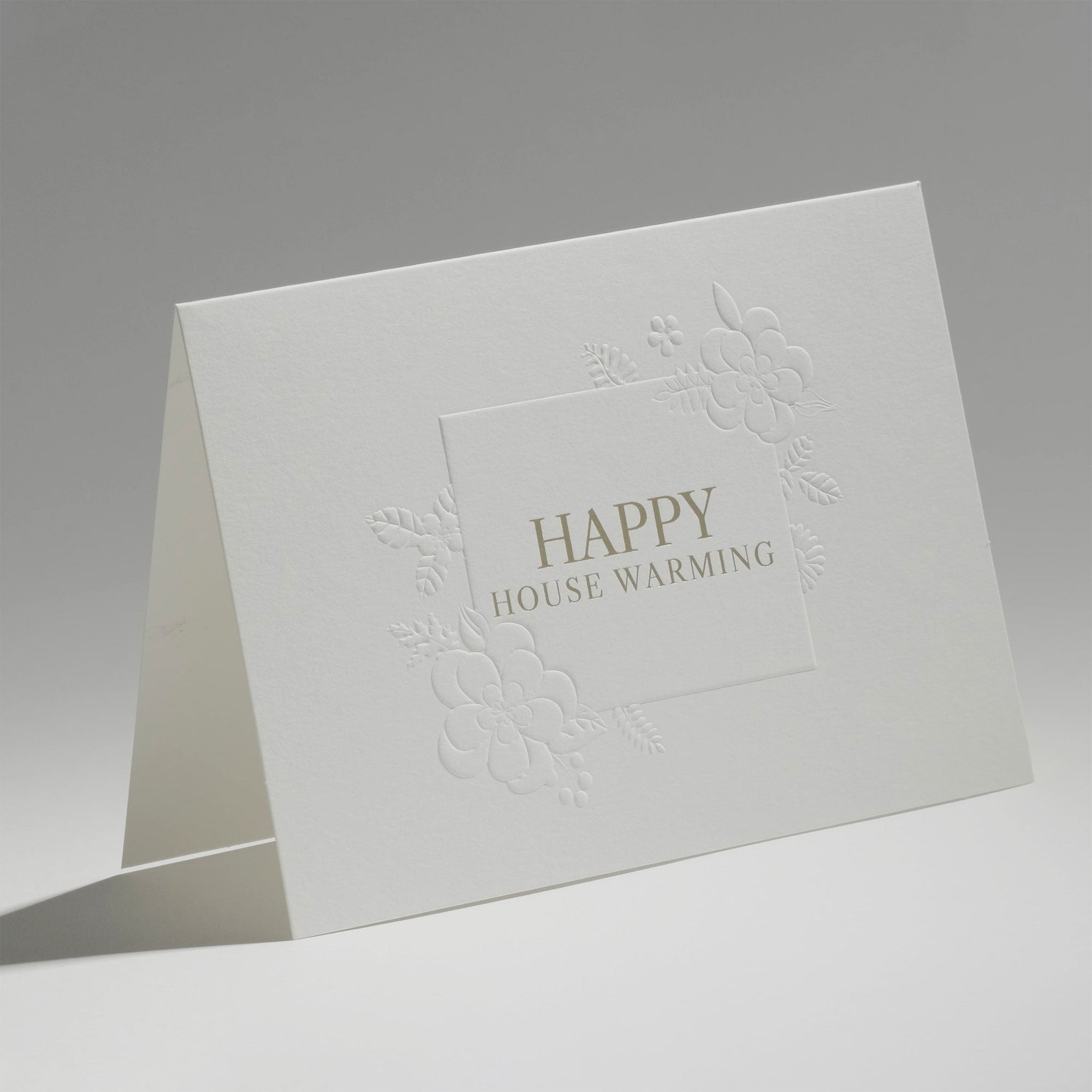 An elegant, freestanding white housewarming greeting card showcasing a detailed blind-embossed floral frame around the central gold typography.