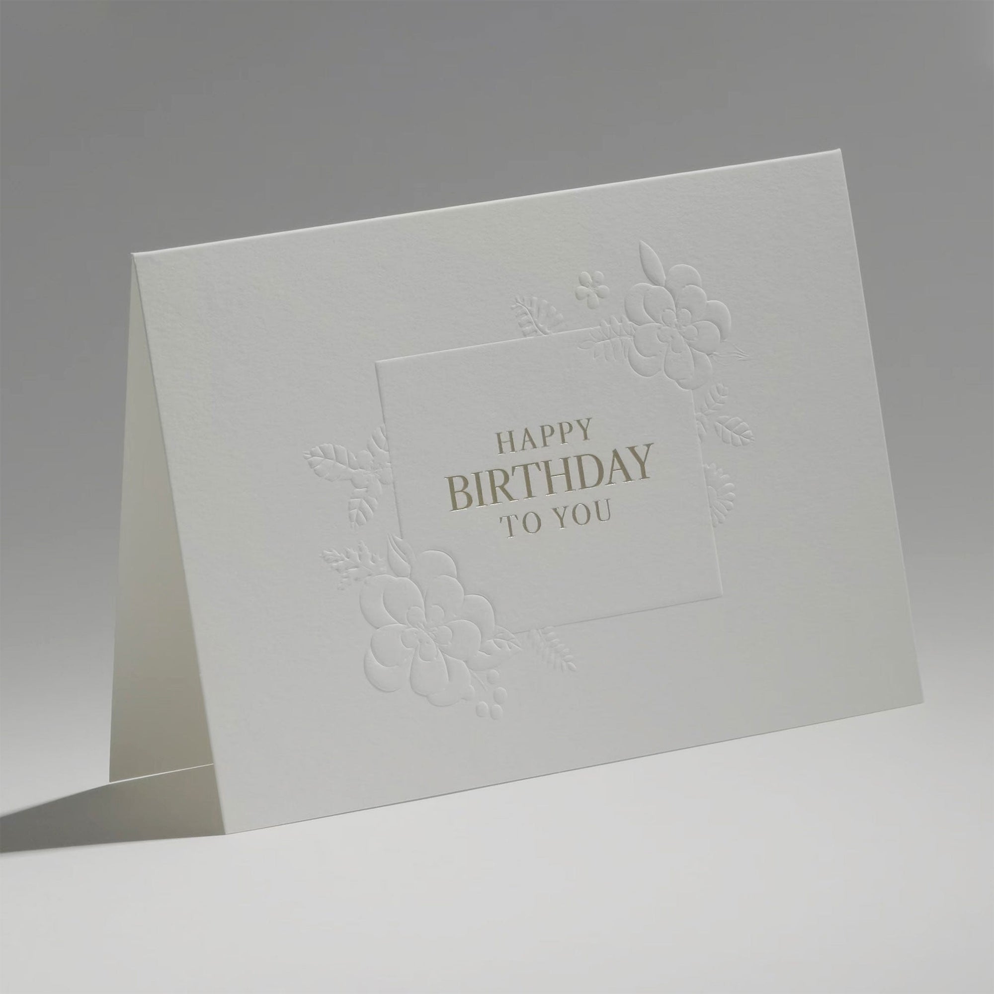 An elegant, freestanding white birthday card displaying a detailed debossed floral border surrounding a clean central birthday greeting.