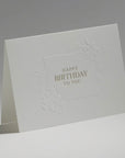An elegant, freestanding white birthday card displaying a detailed debossed floral border surrounding a clean central birthday greeting.