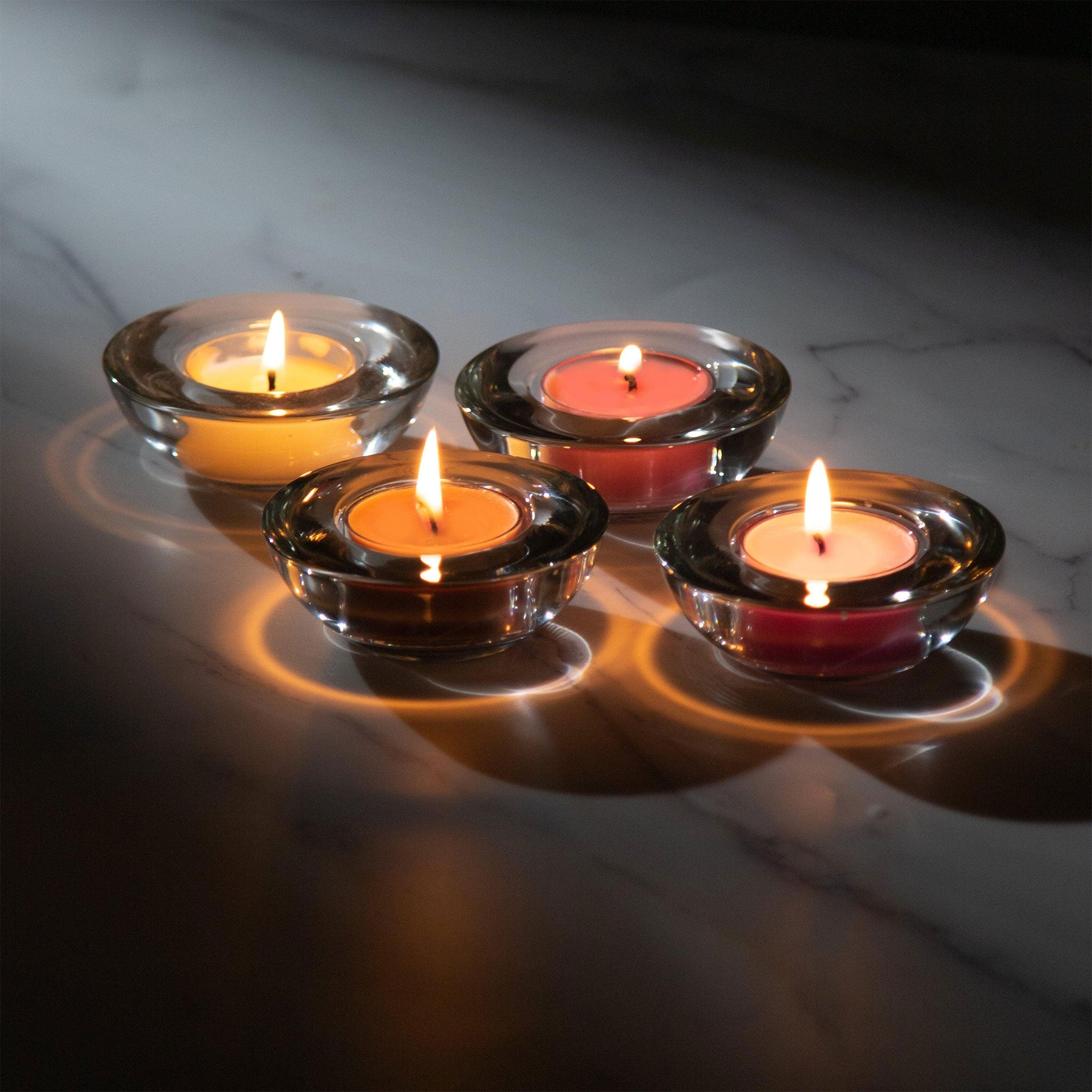 Four lit tealight candles nestled in round glass holders, casting a warm luxury glow and soft circular reflections across a dark surface.