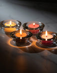 Four lit tealight candles nestled in round glass holders, casting a warm luxury glow and soft circular reflections across a dark surface.