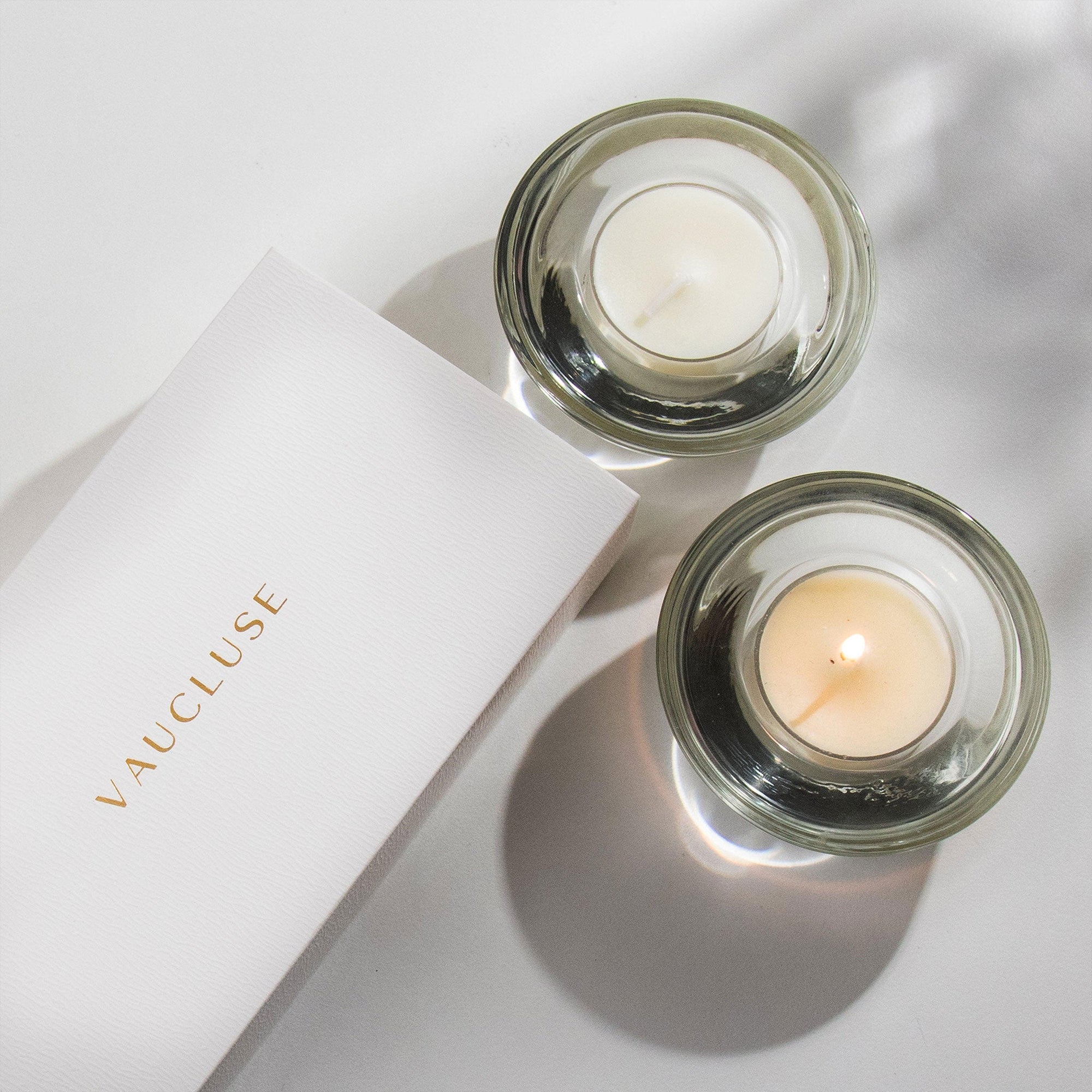 Top down view of two Vaucluse Jasmine scented tealight candles in circular glass holders, one lit with a warm flame, next to a minimalist white brand package.