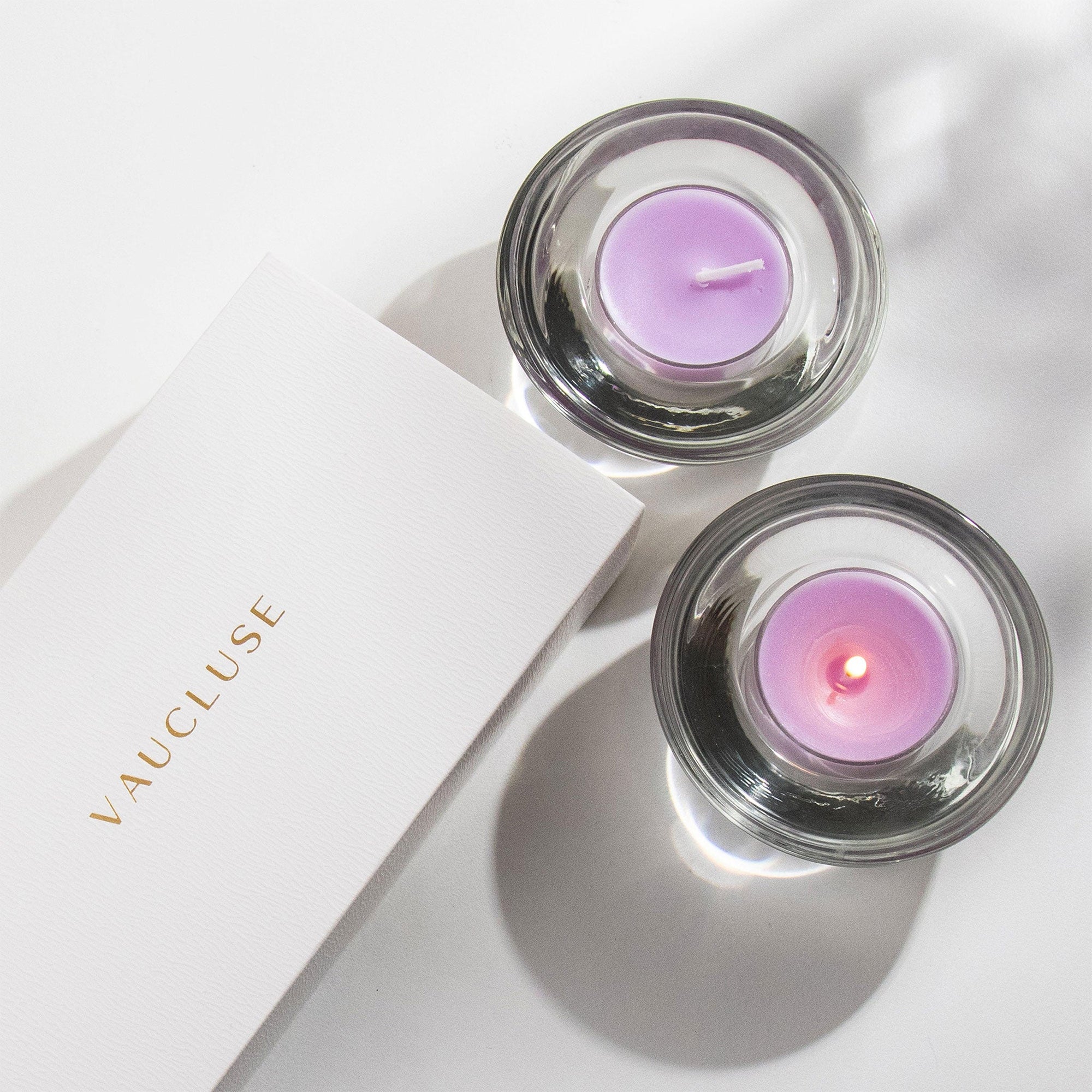 Overhead top down view of two purple Vaucluse Lavender tealight candles in circular glass holders, with one lit and glowing next to a minimalist white brand package.
