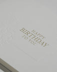 Close-up macro view highlighting the rich, heavy paper texture and crisp gold foil embossing on the Vaucluse "Happy Birthday To You" message.