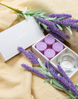 Cozy aromatherapy lifestyle scene featuring the open Vaucluse lavender tealight set resting on draped beige linen fabric with sprigs of lavender.
