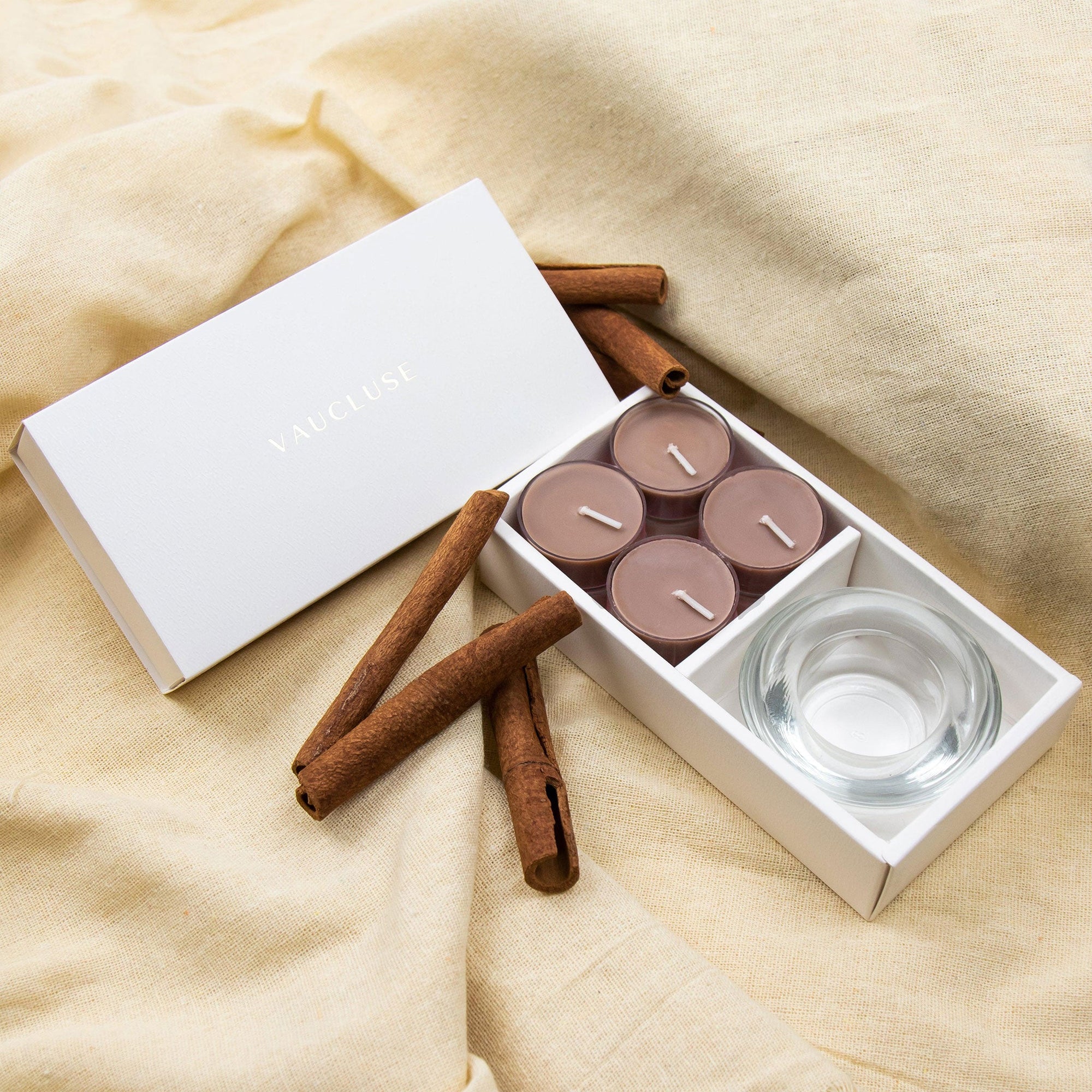 warm lifestyle display featuring the open Vaucluse cinnamon tealight set resting on draped beige linen fabric with whole cinnamon sticks.