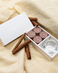 warm lifestyle display featuring the open Vaucluse cinnamon tealight set resting on draped beige linen fabric with whole cinnamon sticks.
