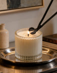 Trimming the wick of a lit luxury white candle in a ribbed glass jar using a matte black candle wick trimmer.