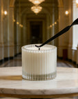 A matte black candle wick dipper straightening the wick of a white candle inside a ribbed glass jar.