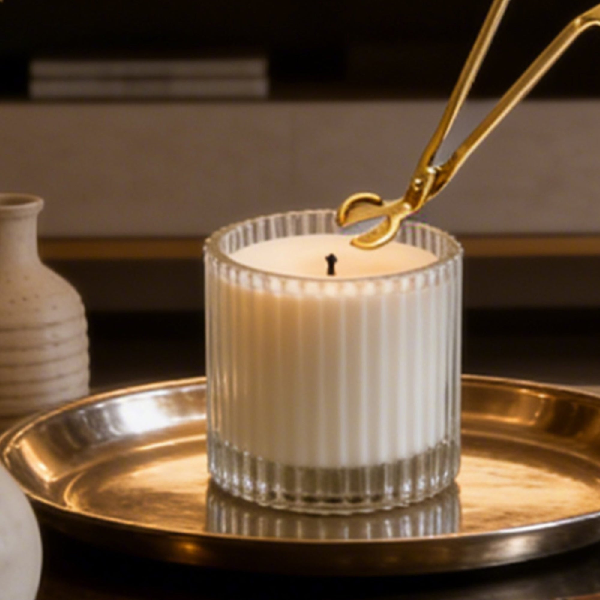 Trimming a candle wick with a polished gold wick trimmer tool on a lit white candle in a ribbed glass jar.