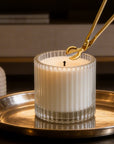 Trimming a candle wick with a polished gold wick trimmer tool on a lit white candle in a ribbed glass jar.