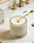 Adjusting a candle wick using a polished gold candle wick dipper inside a bathroom setting.