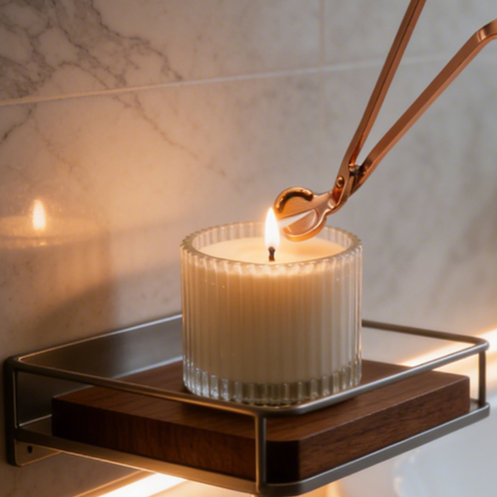 Trimming a candle wick with an elegant rose gold wick trimmer tool on a white candle inside a ribbed glass jar.