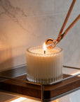 Trimming a candle wick with an elegant rose gold wick trimmer tool on a white candle inside a ribbed glass jar.