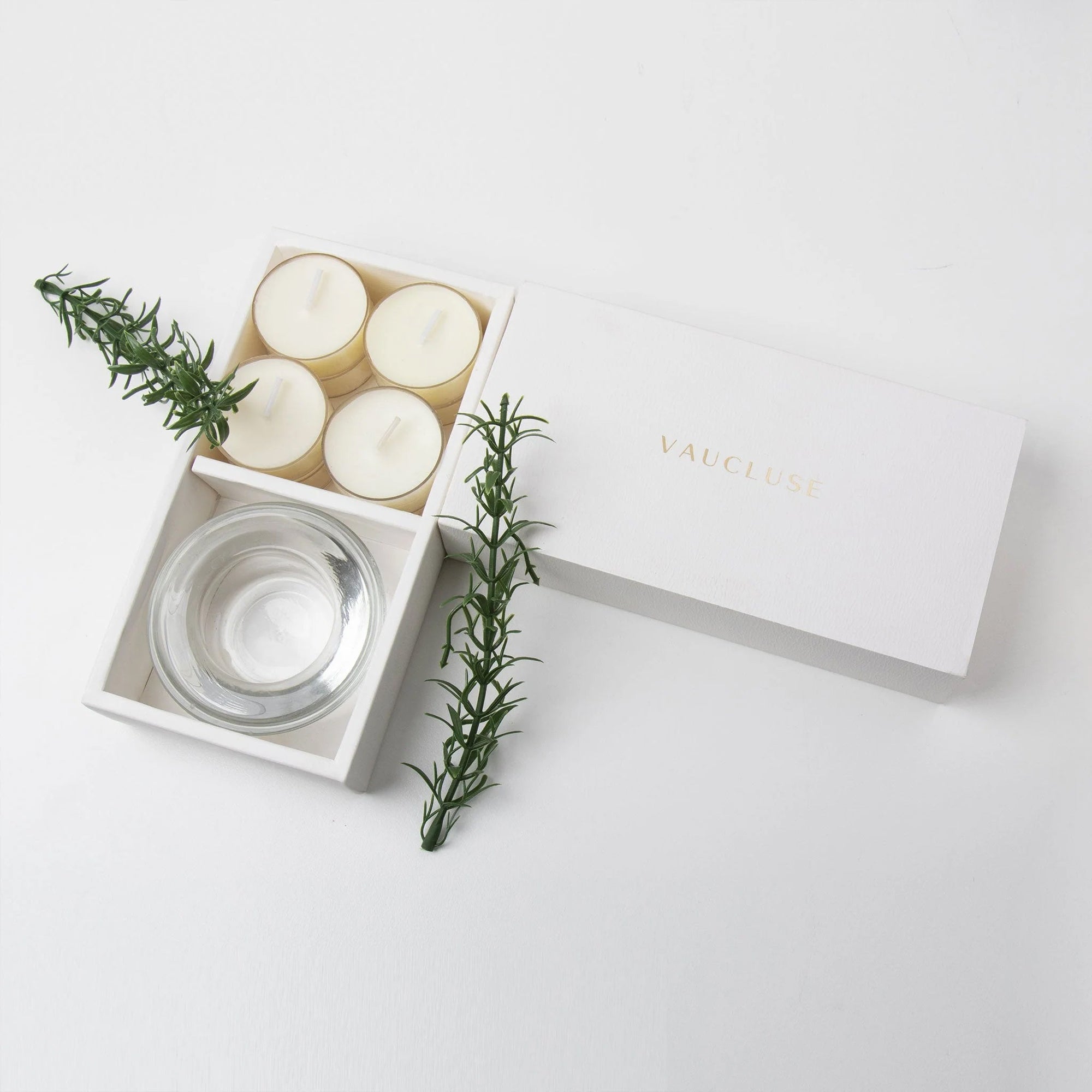 Minimalist top-down view of the open vanilla tealight set and glass candle holder, beautifully styled with fresh rosemary sprigs on a crisp white surface.