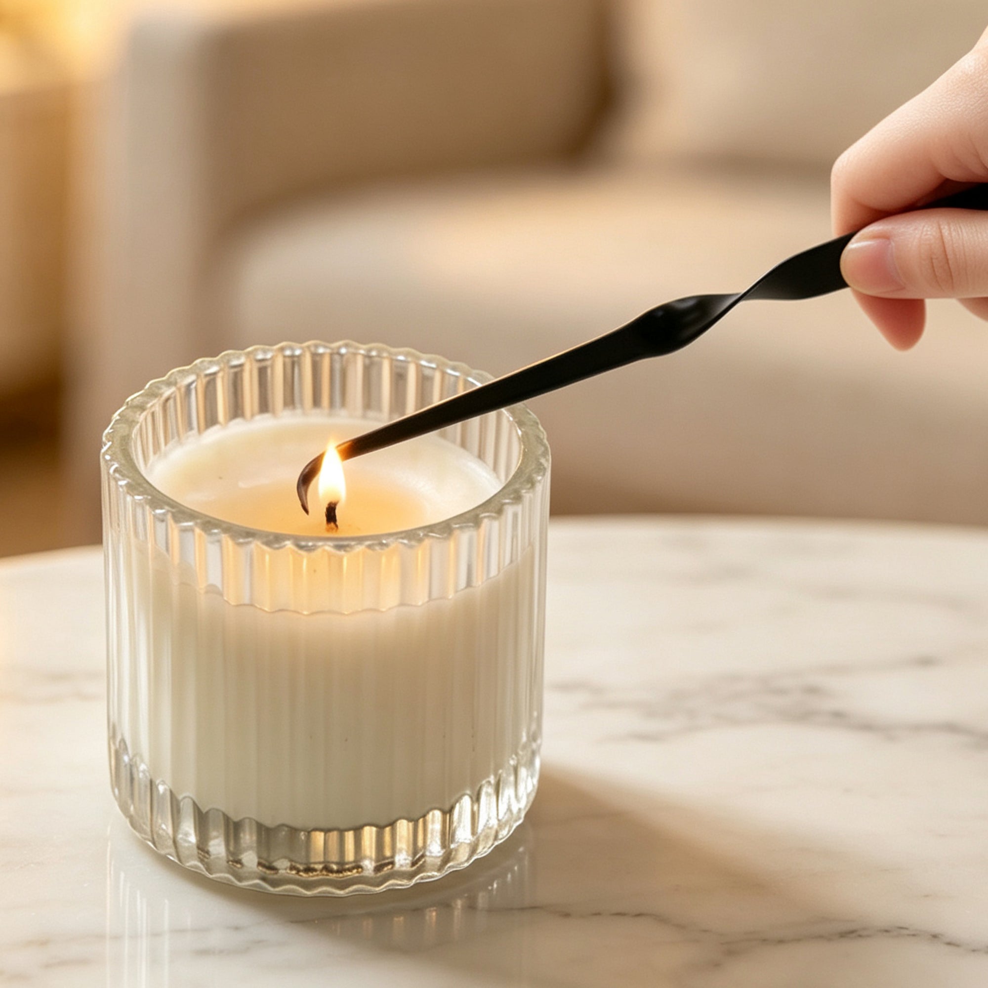 A hand using a twisted black candle wick dipper to adjust the wick of a luxury scented candle in a ribbed glass container.