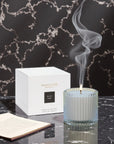 A lit Vaucluse Black Rose scented candle emitting a wisp of smoke, placed next to its minimalist white packaging box on a dark marble surface.