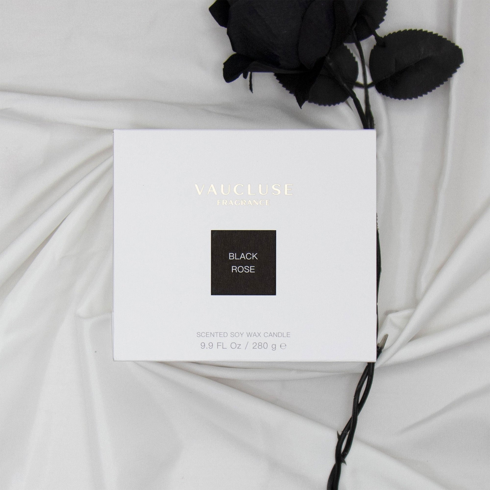 Top down view of the white Vaucluse Black Rose luxury candle gift box resting on soft draped fabric next to a dark black rose stem.